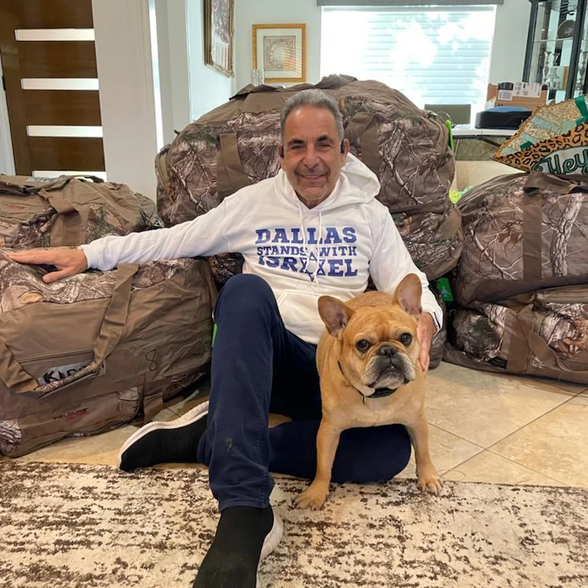 Ivan Sacks sitting on the floor of his Dallas home in a Dallas Stands With Israel sweatshirt, surrounded by packed duffel bags, with his dog beside him