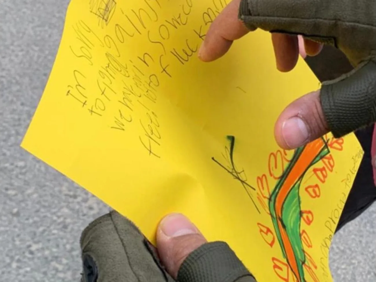 A soldier’s gloved hands holding a bright yellow handwritten letter with a child’s drawing on it