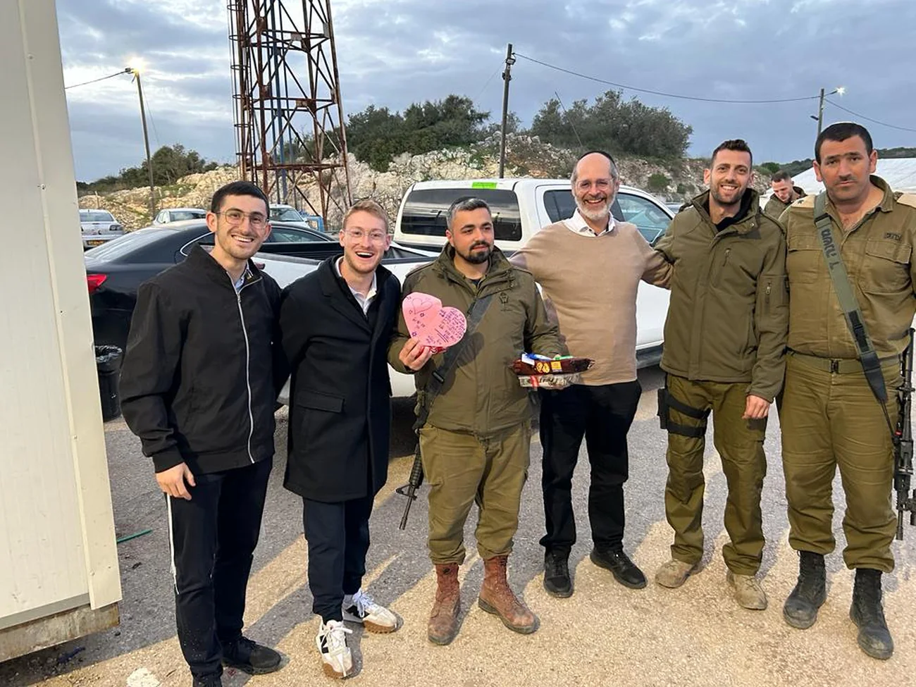 Volunteers and Israeli soldiers standing together beside vehicles, one holding a heart-shaped gift box
